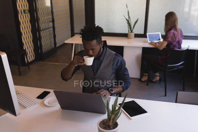 Умный бизнесмен пьет кофе во время использования ноутбука в офисе — стоковое фото