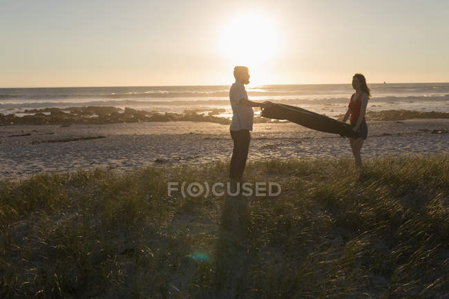 Casal segurando cobertor na praia durante o pôr do sol — Fotografia de Stock