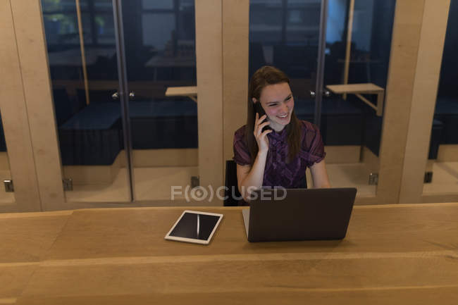 Бізнес-леді розмовляє на мобільному телефоні, використовуючи ноутбук за столом в офісі — стокове фото