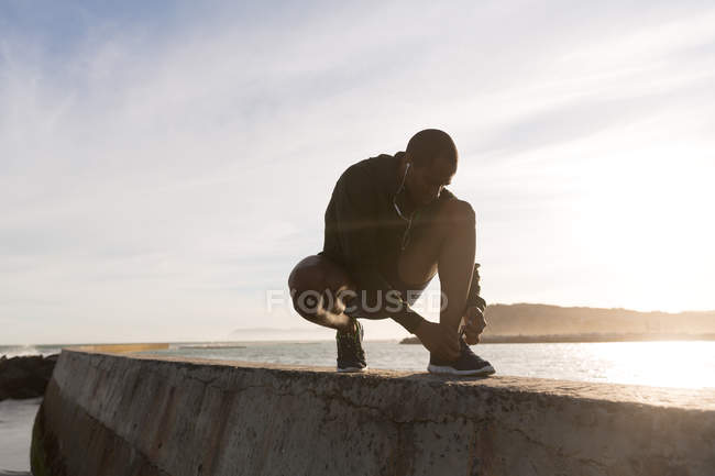 Athlète masculin attachant ses lacets sur le mur environnant à la plage — Photo de stock