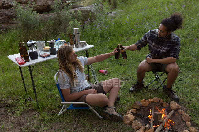 Hombre toasting beer bottle near campfire at camping - foto de stock