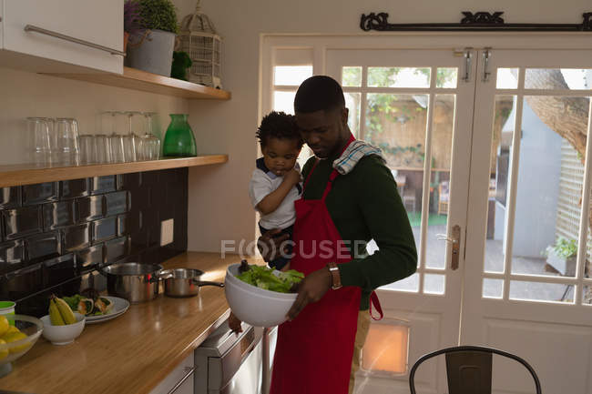 Pai cozinhar comida enquanto segurando seu filho na cozinha em casa — Fotografia de Stock