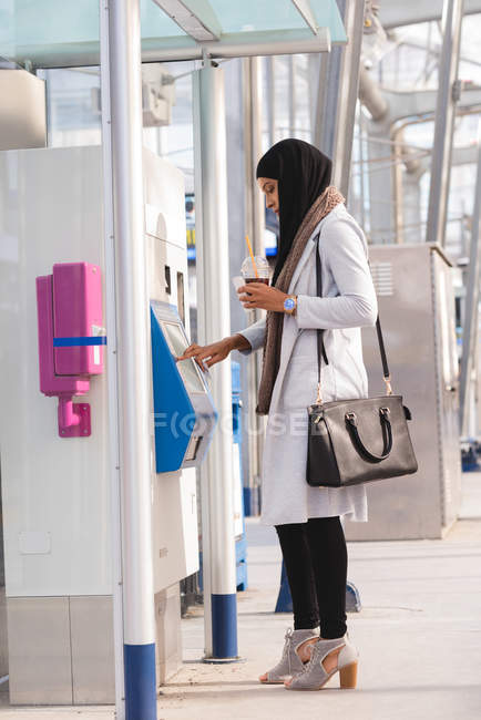 Vista lateral de la mujer hijab utilizando la máquina expendedora de billetes en la estación de tren - foto de stock