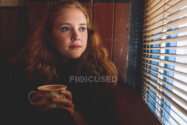 Thoughtful redhead woman looking through window blind in cafe — Stock Photo