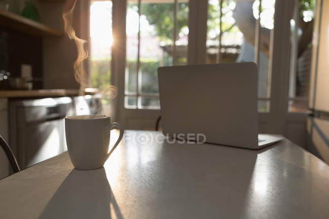 Laptop and cup of tea on table in kitchen at home — Stock Photo