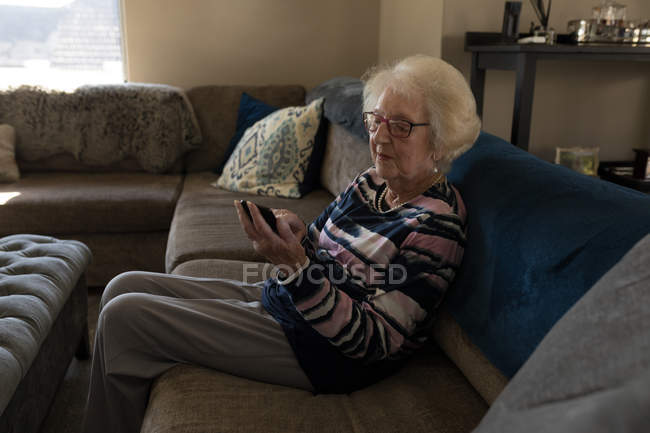 Donna anziana che utilizza il telefono cellulare sul divano in soggiorno a casa — Foto stock