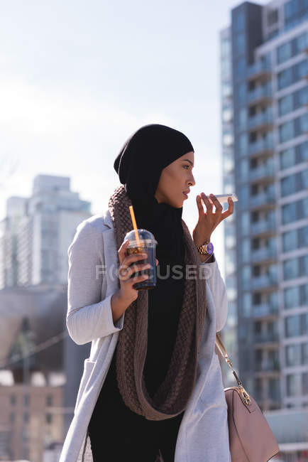 Hijab mujer tomando café frío mientras habla por teléfono móvil en la ciudad - foto de stock