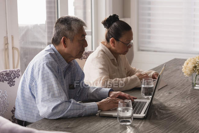 Старшая пара, использующая ноутбук и цифровой планшет на обеденном столе дома — стоковое фото