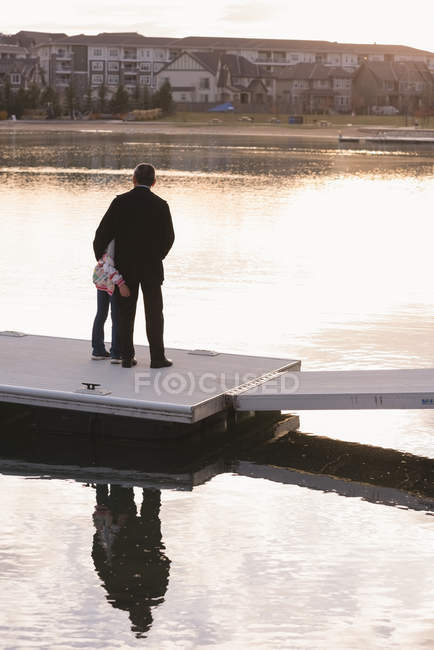 Rear view of grandfather and granddaughter standing on pier near lake side — Stock Photo