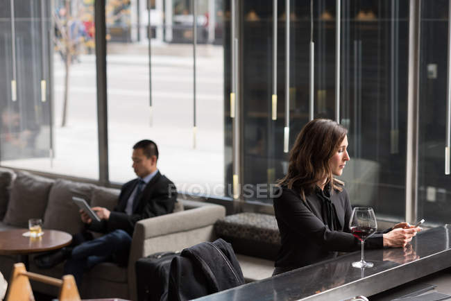 Empresaria usando teléfono móvil en el mostrador del bar mientras hombre de negocios usando tableta digital en el sofá en el hotel - foto de stock