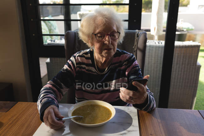 Mulher sênior usando telefone celular enquanto tem comida em casa — Fotografia de Stock
