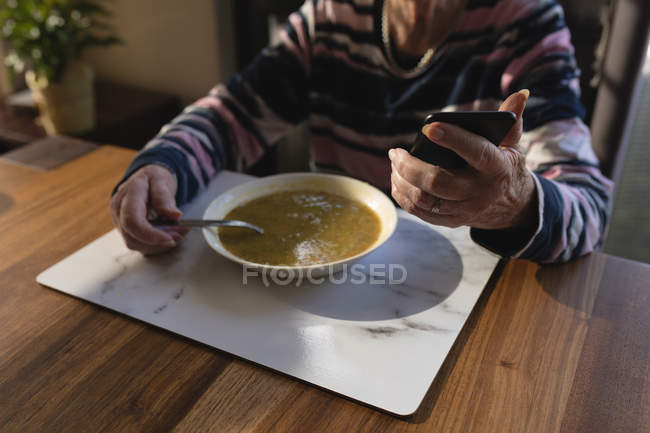 Sección media de la mujer mayor que usa el teléfono móvil mientras tiene comida en casa - foto de stock