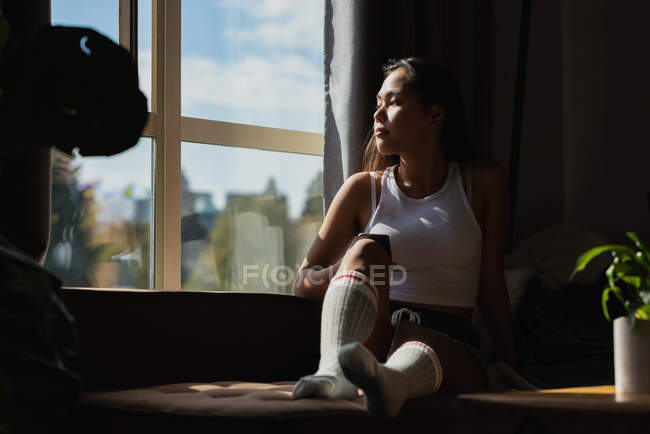 Femme regardant par la fenêtre tout en utilisant le téléphone portable dans le salon à la maison — Photo de stock