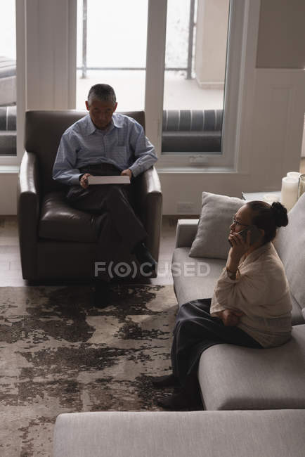 Couple aîné utilisant une tablette numérique et parlant sur un téléphone mobile à la maison — Photo de stock