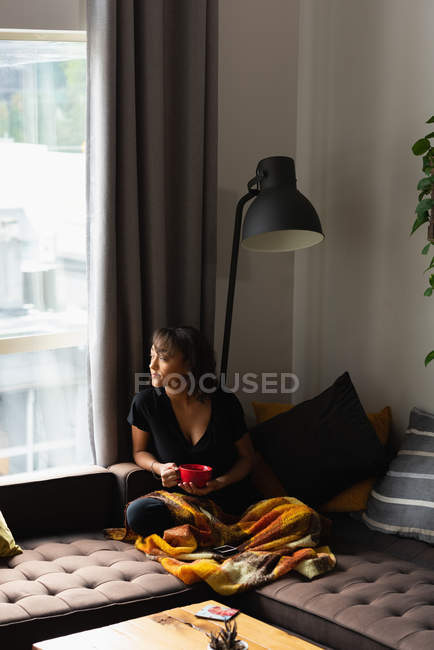 Жінка дивиться крізь вікно, маючи каву на дивані у вітальні вдома — стокове фото