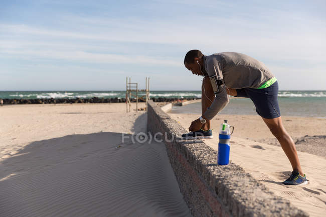 Atleta masculino amarrando seus cadarços na parede circundante na praia — Fotografia de Stock