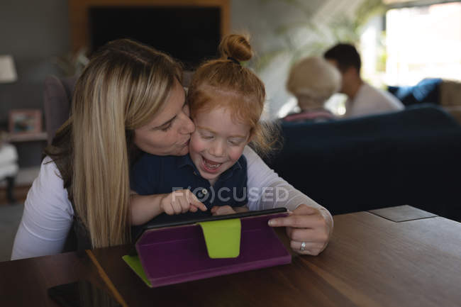 Mother kissing her daughter while using digital tablet in living room at home — Stock Photo