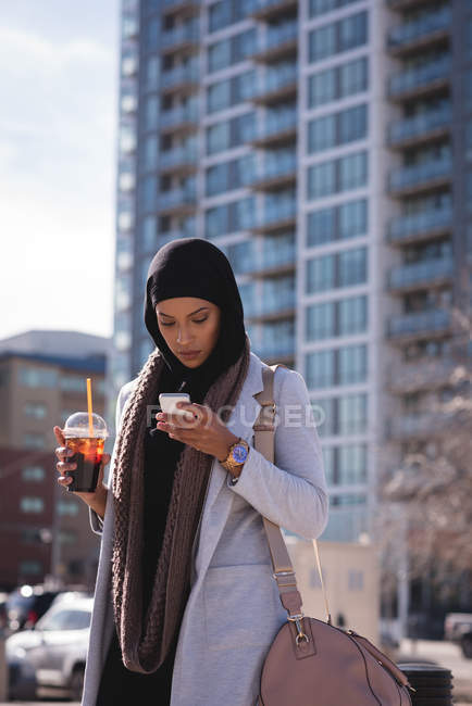 Hijab donna che beve caffè freddo durante l'utilizzo del telefono cellulare in città — Foto stock