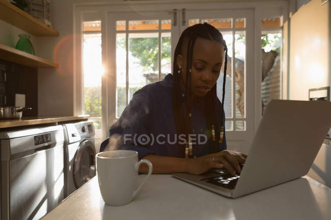 Woman using laptop in kitchen while having coffee on table at home — Stock Photo