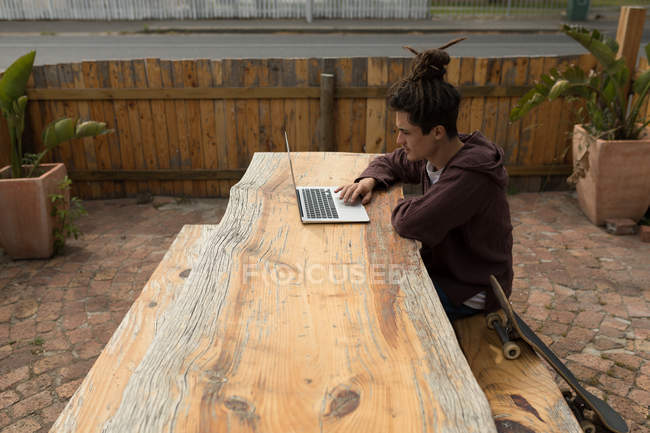 Молодой скейтбордист использует ноутбук в кафе на открытом воздухе — стоковое фото