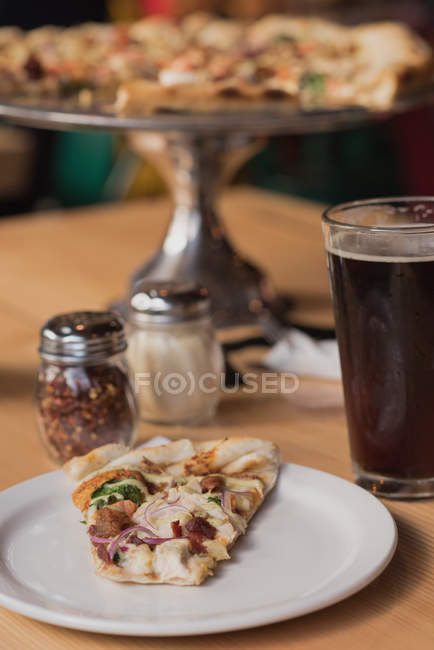 Primer plano de la rebanada de pizza, vaso de cerveza y especias sobre la mesa - foto de stock