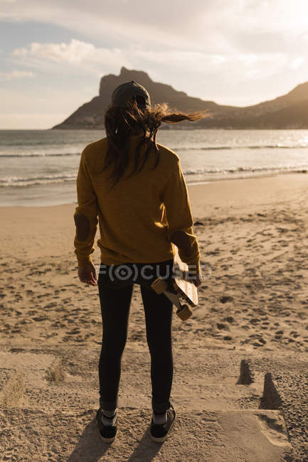 Вид сзади женщины, стоящей со скейтбордом на пляже — стоковое фото