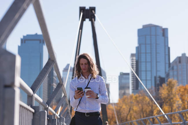 Hombre usando el teléfono móvil mientras toma café en el puente de la ciudad - foto de stock