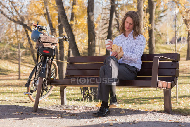 Молодий чоловік має каву, сидячи на лавці в парку — стокове фото