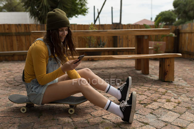 Elegante skatista feminino usando telefone celular enquanto está sentado no skate — Fotografia de Stock