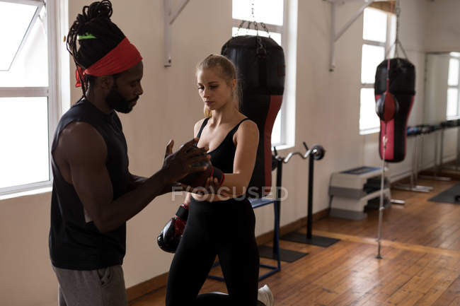 Treinador masculino auxiliando boxeador feminino no uso de luvas de boxe no estúdio de fitness — Fotografia de Stock