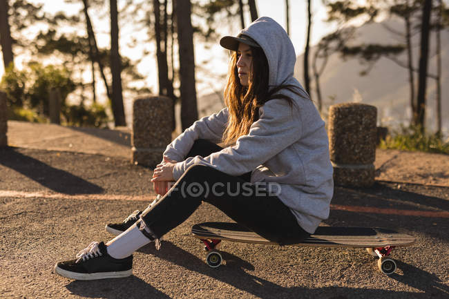 Thoughtful female skateboarder sitting on skateboard at country road — Stock Photo
