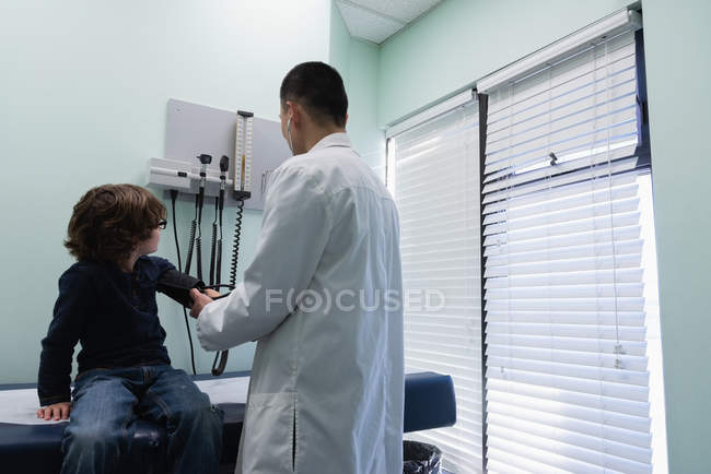 Rear view of young asian male doctor checking blood pressure of caucasian boy patient in clinic. Male doctor using sphygmomanometer — Stock Photo