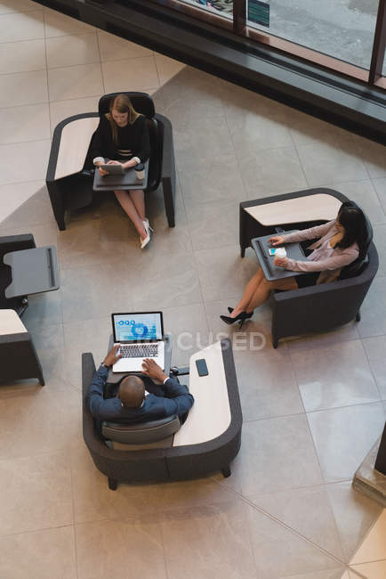 Vista superior de gente de negocios usando dispositivos multimedia sentados en los sillones en el vestíbulo en la oficina - foto de stock