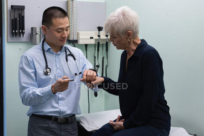 Молодой врач-азиат измеряет уровень сахара в крови с помощью глюкометра пожилой женщины в клинике — стоковое фото