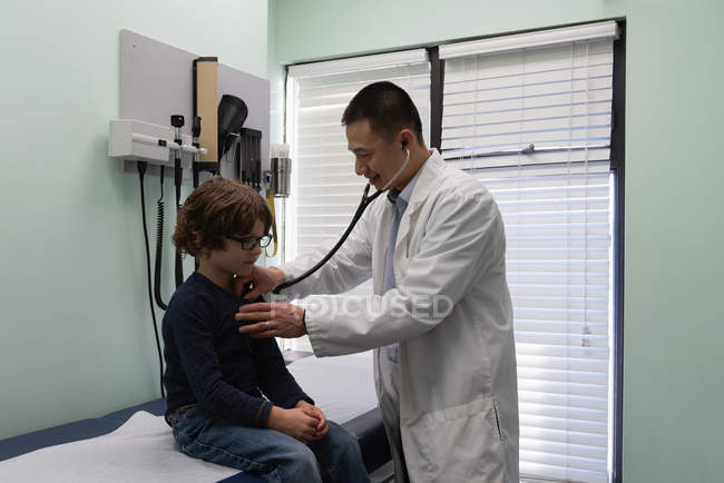 Seitenansicht eines jungen asiatischen männlichen Arztes, der die Brust eines Jungen in einer Klinik untersucht. Arzt mit Stethoskop — Stockfoto