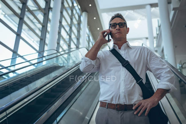Vue de face d'un homme d'affaires parlant sur le téléphone portable tout en descendant sur l'escalier roulant dans un bureau moderne — Photo de stock