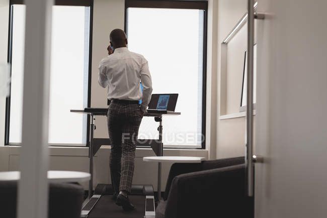 Вид ззаду бізнесмена, який розмовляє по мобільному телефону під час тренувань на біговій доріжці в сучасному офісі — стокове фото