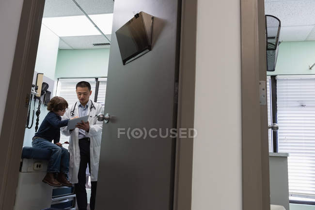 Низкий угол обзора молодого врача-азиата и пациента-белого мальчика с помощью цифрового планшета в клинике. Мужской врач взаимодействует с пациентом — стоковое фото