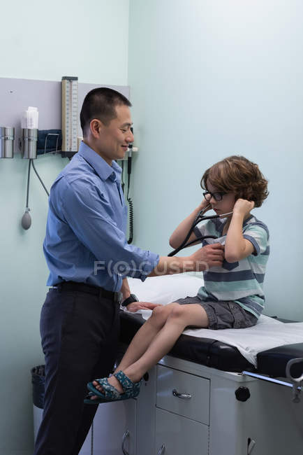 Вид сбоку на молодого врача-азиата, осматривающего пациента белого мальчика со стетоскопом в клинике — стоковое фото