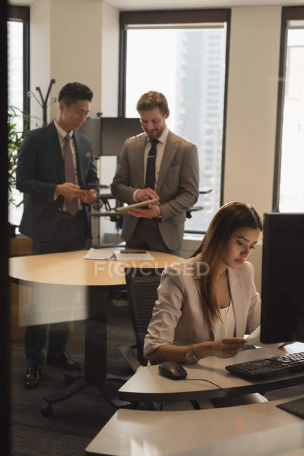 Vista lateral da empresária olhando para o documento enquanto colegas de trabalho discutindo sobre tablet digital no escritório moderno. A mulher está sentada e os homens de pé — Fotografia de Stock