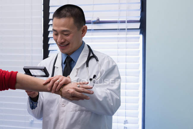 Vista frontal de un joven médico asiático examinando a un paciente mayor con lupa en la clínica - foto de stock