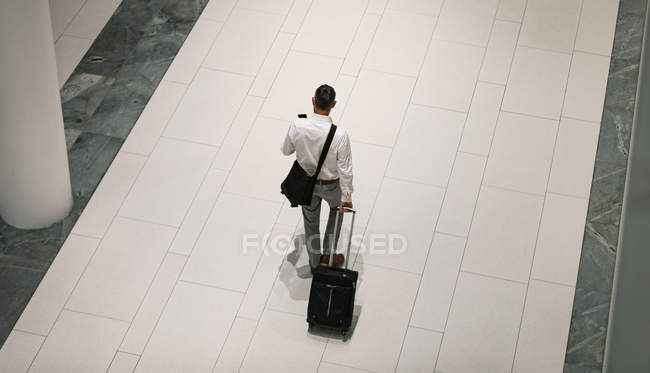 Erhöhte Rückansicht des Geschäftsmannes mit Reisekoffer, der auf sein Handy schaut, während er auf dem Flur geht — Stockfoto