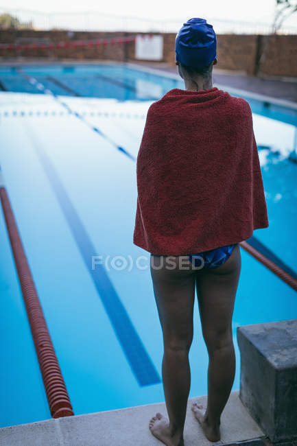 Vista posteriore del giovane nuotatore femminile avvolto in asciugamano in piedi vicino alla piscina — Foto stock