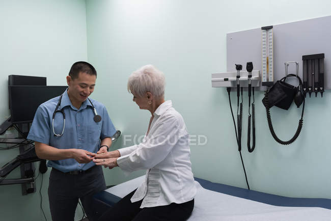 Молодой врач-азиат осматривает пожилую пациентку в клинике, дотрагиваясь до рук — стоковое фото