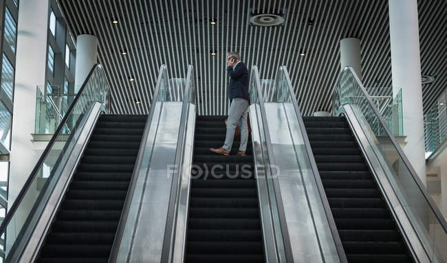 Низкий взгляд бизнесмена разговаривающего по мобильному телефону на эскалаторе в офисе — стоковое фото