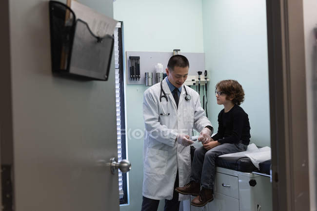 Vista frontal de joven asiático médico masculino examinando caucásico niño paciente con glucosímetro en clínica - foto de stock
