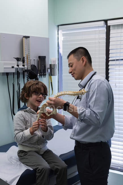 Vue latérale du jeune médecin masculin asiatique montrant le modèle de colonne vertébrale au patient garçon caucasien — Photo de stock