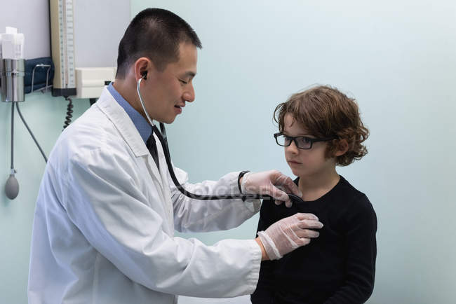 Visão lateral de jovem asiático médico masculino examinando caucasiano menino paciente com ferramenta em uma clínica — Fotografia de Stock