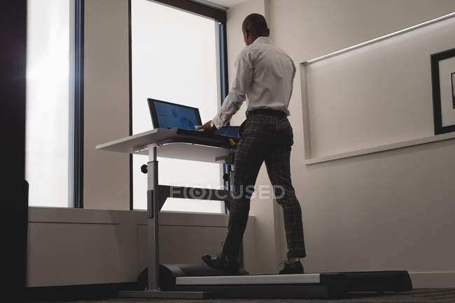 Vista laterale dell'uomo d'affari che utilizza il computer portatile mentre si esercita sul tapis roulant nell'ufficio moderno — Foto stock