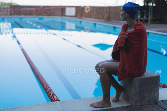 Vista lateral de una joven nadadora envuelta en una toalla sentada cerca de la piscina - foto de stock